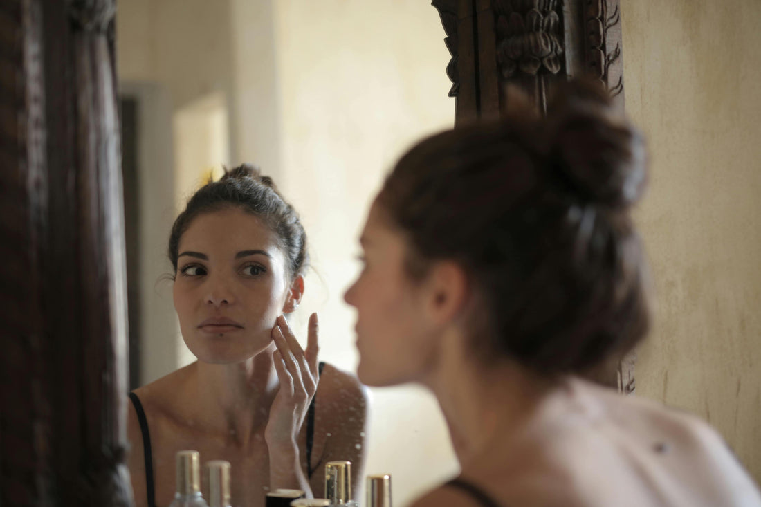 A woman with short brown hair and freckles is touching her face with both hands. She is looking directly at the mirror, assessing and feeling her skin to see how it is feeling and what it needs